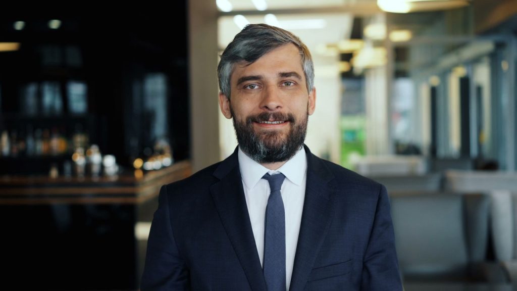 Business professional with a beard smiling confidently in a modern office environment.